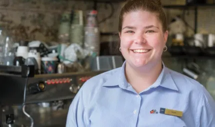 Woman with hair tied back wearing blue shirt stands in front of coffee making machine with cups and shelving in the background.