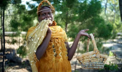 An Indigenous woman models a golden yellow silk dress, scarf and headpiece, holding up in one hand a handwoven-coiled basket.  She is standing in a natural environment, shaded by green trees.