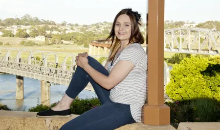 Young woman sits on a wall with her back to a pillar. She is dressed in dark slacks and light shirt. In the background is a tree, a river, two bridges and houses.