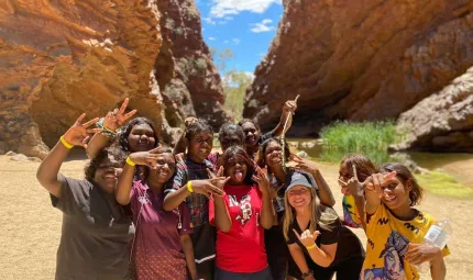 Group of 9 young women from Arlparra, 8 of them Aboriginal, standing proud and happy, close together, posing for the photo in front of a canyon.