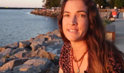 A head and shoulders photo of a woman with long brown hair and necklaces. In the background is water, rocks lining the water, trees and a footpath.