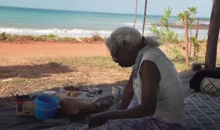 An elderly woman with grey hair sits on a mat with painting equipment in front of her. In the background is red soil, grass, a shrub and the ocean.