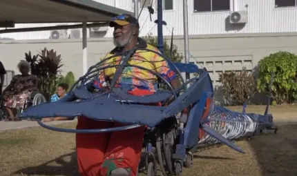 An elderly man sits in a wheel chair that forms part of a long metal contraption in the shape of a hammerhead shark. In the background are two women, grass, shrubbery and a building.