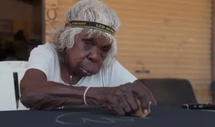 An elderly Aboriginal woman wearing a headband around her grey hair sits at a table and sketches on a black canvas.