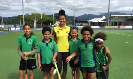 A young adult Indigenous woman in yellow shirt and black pants stands between five Indigenous children dressed in green shirts and black shorts. All stand on a flat green playing surface. In the background are poles, and buildings.