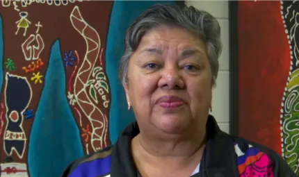 A woman sits in front of two brightly coloured Indigenous artworks. She is wearing a black and brightly coloured polo shirt.