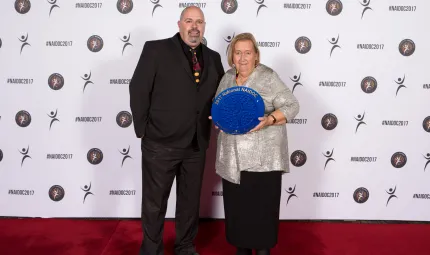 Indigenous male stands next to an older Indigenous woman on red carpet, the woman is holding large blue plate.