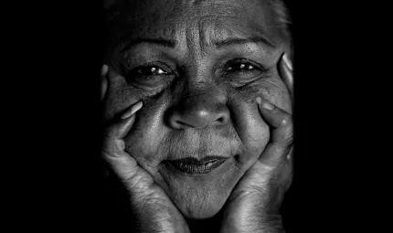 Close up image of Aboriginal woman with face in hands, mouth closed and staring directly into camera.