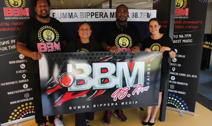 Two Indigenous men and two Indigenous women in black t-shirts stand in a room on a hard floor and behind a banner which says BBM 98.7FM. Behind them are black vertical banners with similar logos and writing.