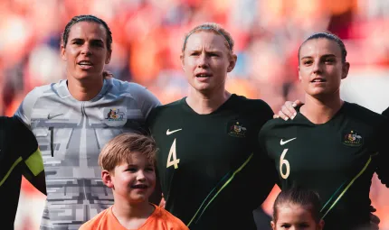 Three women (one in grey shirt and two in dark green shirts) stand arm in arm with two children in front of them. In the background is a crowd of people mainly in orange clothing.