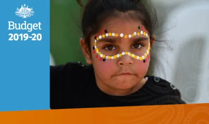 A primary-school aged girl looks at the camera. She is wearing a black and white striped jumper. She is wearing yellow, white, red and black face paint in the traditional dot style around her eyes.