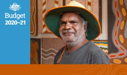 A man wearing a grey shirt and a wide brimmed straw hat is standing in front of Indigenous artwork comprised of wavy and straight lines and red and yellow large dots. Beside there is a blue rectangle with Budget 2020 – 2021 and the Australian Government C