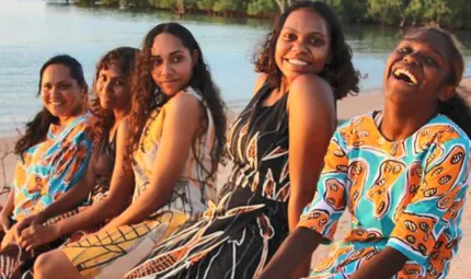 Five Indigenous women wearing colourful clothing sit in a line on a beach with water and trees in the background.