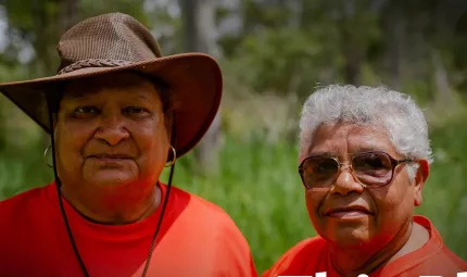 Two Aboriginal women in crimson t-shirts, one wearing a brown brimmed hat and the other with grey hair and wearing sunglasses, look to camera. In the background is green grass and trees.