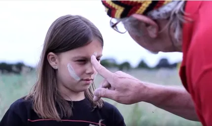 The face of a young girl is painted with white stripes by an elderly man in red shirt and head wear in black, red and yellow.