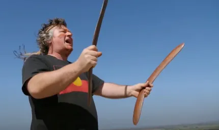 A middle aged man with wavy hair dressed in black shirt with Aboriginal flag on the front holds a boomerang in each hand and is in the attitude of singing as he looks out across the landscape. In the background is blue sky and a strip of green land.