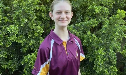 Young woman in maroon shirt with hair tied back stands in front of a green shrub.