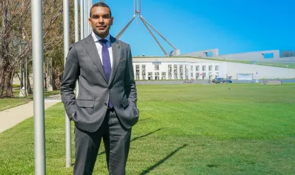 Image of a man wearing a grey suit, standing on the left, facing the camera with Parliament House behind him, a very blue sky and green grass.