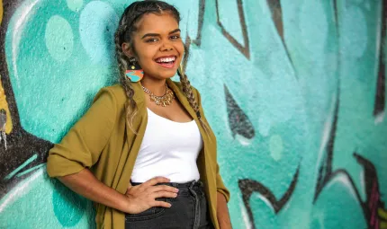 An Indigenous young adult woman, her hair plaited and wearing a green yellow cardigan, white top  and black jeans, leans up against a wall painted black and with an aqua coloured mural.