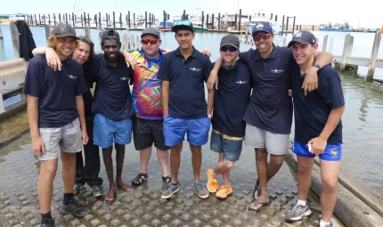 Seven young Indigenous males and one older male, dressed in shorts, shirts and hats. They are standing on a boat ramp with water lapping at their feet. In the background is a dock with boats tied up.