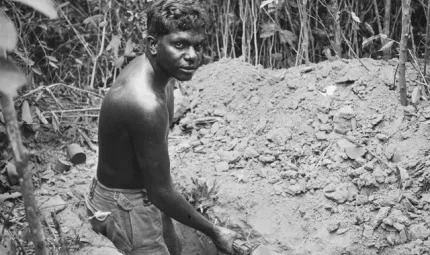 A young man wearing shorts but bare chested holds a shovel in his hand while he stands in a trench. In front of him is a pile of soil and beyond that is thick foliage.