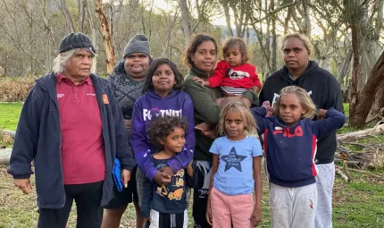 Group of Aboriginal women and children dressed in casual wear stand on a grassy surface. In the background are trees and a hill.