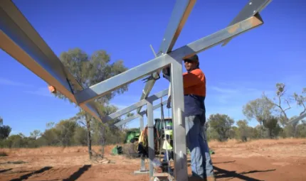 Aboriginal man dressed in workwear, hat and sunglasses works on a steel structure set on red earth with grass and trees in the background and a bright blue sky overhead.