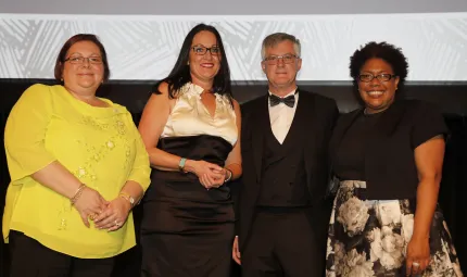 Four adults stand in a row: woman in yellow top, woman in white and black dress, man in tuxedo and woman in black top and black and white mottled skirt. All wear glasses and stand in front of a two toned wall.