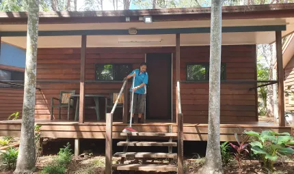 A woman in work wear sweeps the deck in front of a wooden cabin. At the front are two palm trees, some foliage and steps leading up to the cabin entrance. In the background are more trees.