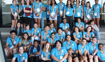 Large group of young Indigenous women sit and stand on several rows of stairs. Most wear a blue shirt.