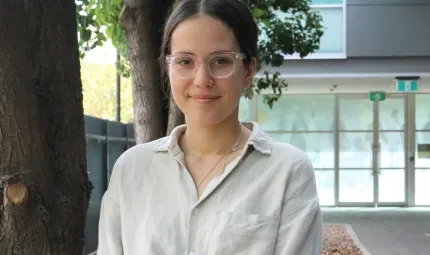 A young woman with dark hair, wearing glasses and a pale shirt stands in front of some trees. In the background and to the right is a building entrance made mainly of glass windows set in metal frames.