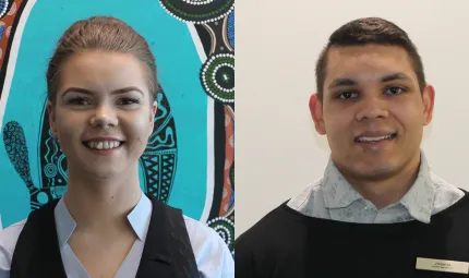 Woman in pale shirt and dark vest standing in front of Indigenous artwork featuring blue, green, black and ochre coloured elements. A man is to her left dressed in pale shirt and dark jumper standing in front of pale, featureless background.