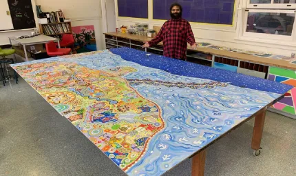 A man wearing a black and red checked shirt is standing in front of a large colourful canvas painting which is on a table.