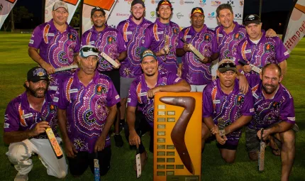 Group of men in purple jerseys featuring Indigenous designs, stand or kneel around a large trophy featuring a boomerang on a large wooden plaque with small metal plates.