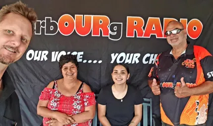 Two women sit in front of a black background with two men standing either side of them. Words on the background are Cherbourg Radio Our Voice…Your CH