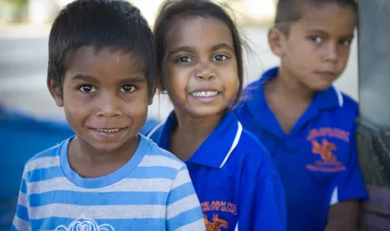 Young Indigenous male child in white and blue striped shirt in front of and to left of similar aged Indigenous girl in dark blue shirt who is in front of another Indigenous boy in same type of shirt.