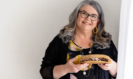 A woman – head tilted to her left, and bearing a broad, friendly smile – cradles an array of chocolates in a coolamon (a traditional Indigenous carrying vessel). She is wearing clothing and jewellery in a mixed palette of bright earth and dark tones.