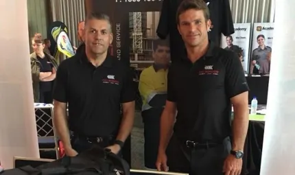 Two men in dark shirts stand facing camera. In the background are posters and a woman with arms folded.