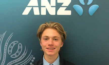 Young man with blonde hair wearing dark jacket, vest and tie stands below a sign which says ANZ.