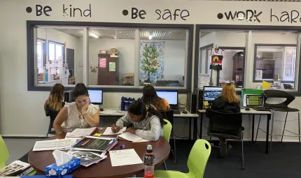 Four young women and an adult woman sit at computer desks or a table in a carpeted room with pale coloured walls and windows through to another room. Above the windows are the words: Be kind, Be safe, Work hard.