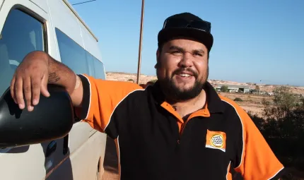 Indigenous man leaning up against a white van with buildings in the background.