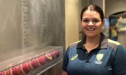 Aboriginal woman in dark blue shirt stands next to a glass display cabinet featuring red cricket balls.