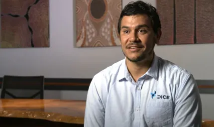 A bearded man in a pale shirt sits in front of a large wooden table. In the background are paintings of traditional design.