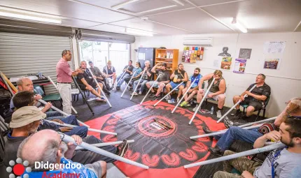 A group of men sit in a circle on chairs in a well-lit room. Each holds a white pipe pointed at the floor on which is a red and black covering. On the walls are images, a book shelf and other items. There are two roller doors on one wall.