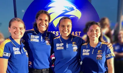 Four Indigenous women in blues shirts stand shoulder to shoulder. They wear blue and yellow shirts. Behind them is a sign with a logo featuring an eagle’s head.