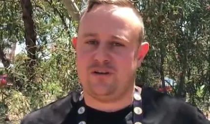 Indigenous man in black shirt stands in front of trees