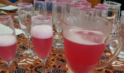 Several glasses and a jug sit on a tablecloth of Indigenous design. Some glass are empty while the jug and other glasses are filled with a red drink with a white head.