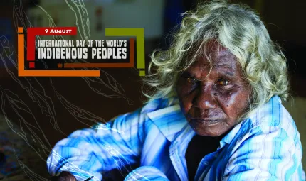 White haired Aboriginal woman wearing blue and white collared shirt sits on a floor. To her left is a graphic of leaves and the following words: 9 August International Day of the World’s Indigenous Peoples.