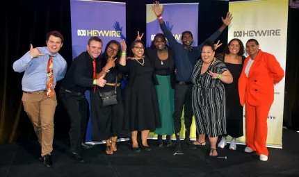 Group of 9 casually dressed Indigenous young adults stand on a black floor in front of three banners which display a logo and the word Heywire.