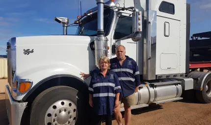 A woman and a man in blue and white work wear stand on brown soil in front of a large white truck. In the background is blue sky.